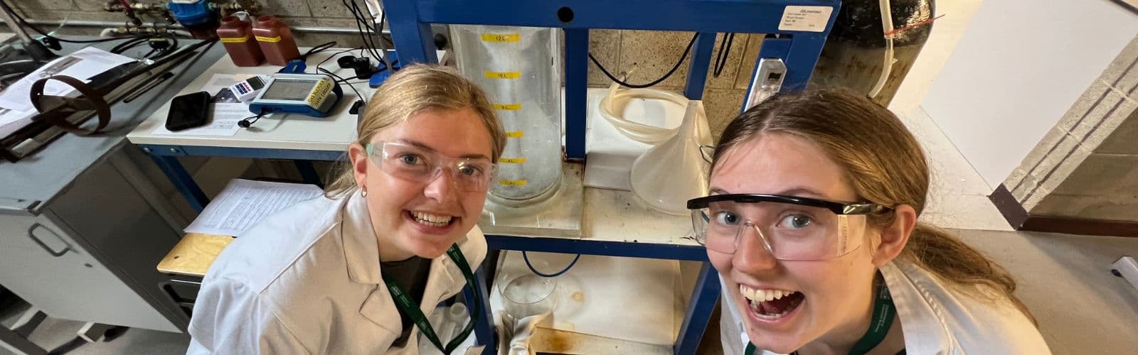 Two students wearing lab coats and goggles pose in front of a lab station. 